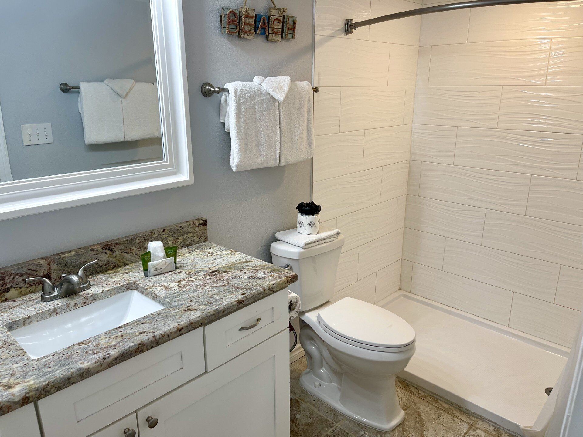 A bathroom with a granite countertop sink, white cabinets, a toilet, and a shower area.