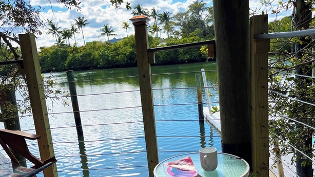Una terraza de madera con una mesa y sillas de vidrio con vista a un cuerpo de agua tranquilo, rodeada de exuberante follaje.