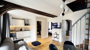 A modern living room with a sofa, a glass coffee table, and a television. There is a kitchen area with white cabinets and a marble countertop. The room has wooden beams on the ceiling and a staircase with metal railings.