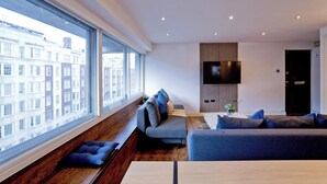 A modern living room with a large window, a grey sofa, a wooden bench, and a flat-screen TV mounted on the wall. - Imperial Queensway Apartment (London)