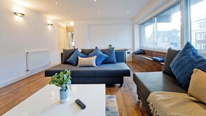A modern living room with a dark sofa set, a white coffee table with a remote control, and a large window offering a view of buildings. - Imperial Queensway Apartment (London)