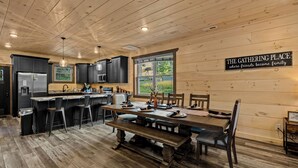 Une cuisine spacieuse avec un plafond en bois, un coin repas avec une longue table et des chaises, et une inscription sur le mur qui dit : « L’ENDROIT OÙ L’ON SE RASSEMBLE, là où les amis deviennent une famille. »