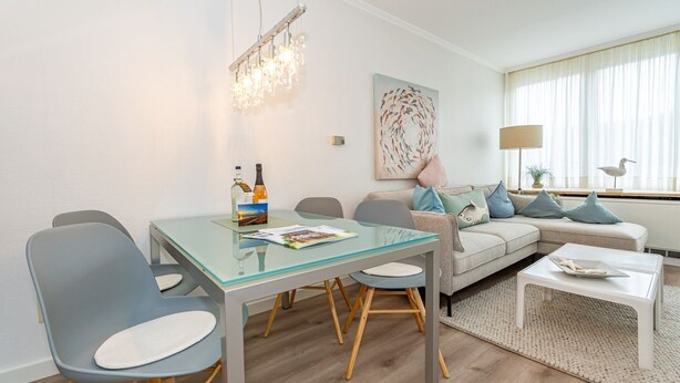 Ein modernes Wohnzimmer mit einem Glastisch, grauen Stühlen, einem grauen Sofa mit blauen Kissen und einem weißen Couchtisch sowie einer Kristallleuchter.