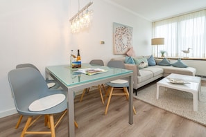 A modern living room with a glass dining table, grey chairs, a grey sofa with blue cushions, a white coffee table, and a chandelier. - Lieblingsplatz - Comfortable vacation apartment Lieblingsplatz with balcony and (Sylt)