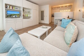 A modern living room with a sofa, a coffee table, and a TV on a stand. There are bookshelves and a kitchen area visible in the background. - Lieblingsplatz - Comfortable vacation apartment Lieblingsplatz with balcony and (Sylt)