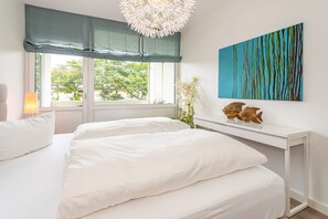 Une chambre avec un grand lit, un bureau blanc, un tableau accroché au mur et une fenêtre donnant sur des arbres.