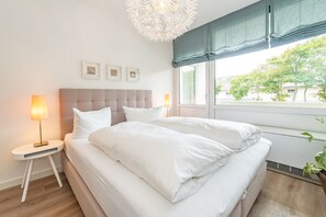 A neatly made bed with white linens, a bedside lamp, and a window with a view of greenery outside. - Lieblingsplatz - Comfortable vacation apartment Lieblingsplatz with balcony and (Sylt)