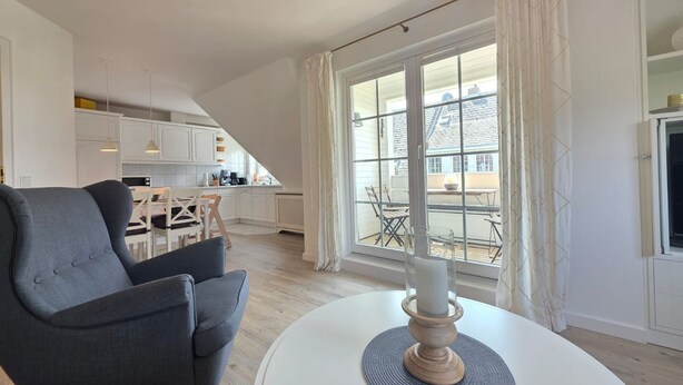 Een moderne woonkamer met een grijze fauteuil, een ronde witte tafel met een kaars, en uitzicht op een balkon met een eettafel en stoelen.