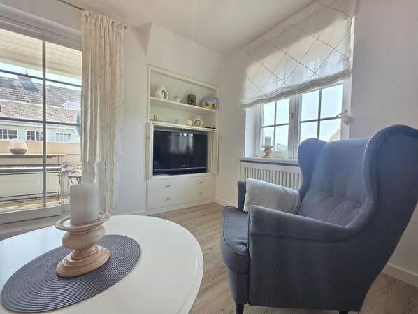 Una sala de estar con televisión, un sillón azul, una mesa blanca con una vela y vistas a edificios.