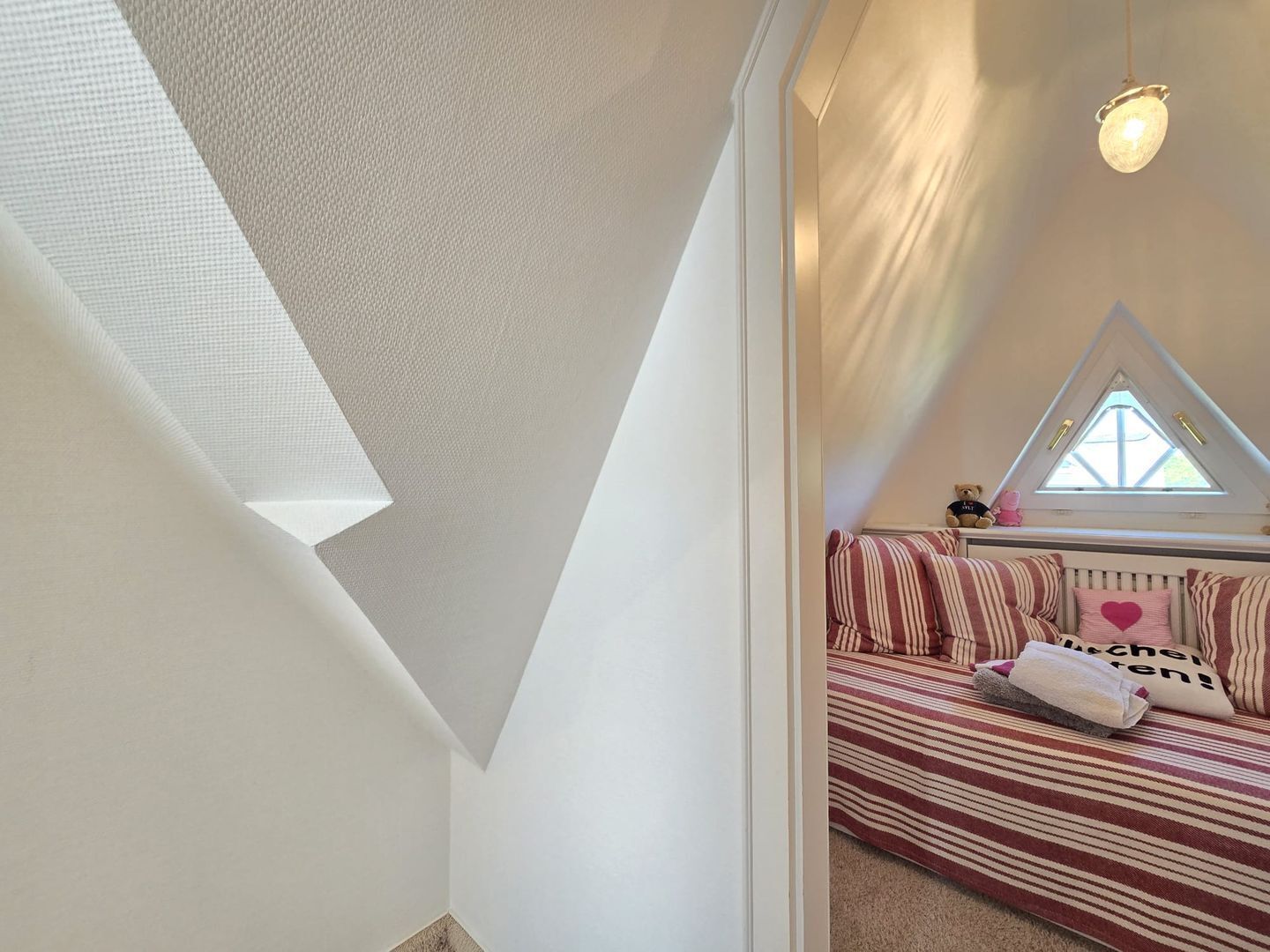 A bedroom with a sloped ceiling, a window, a bed with striped bedding, and a teddy bear on the windowsill.