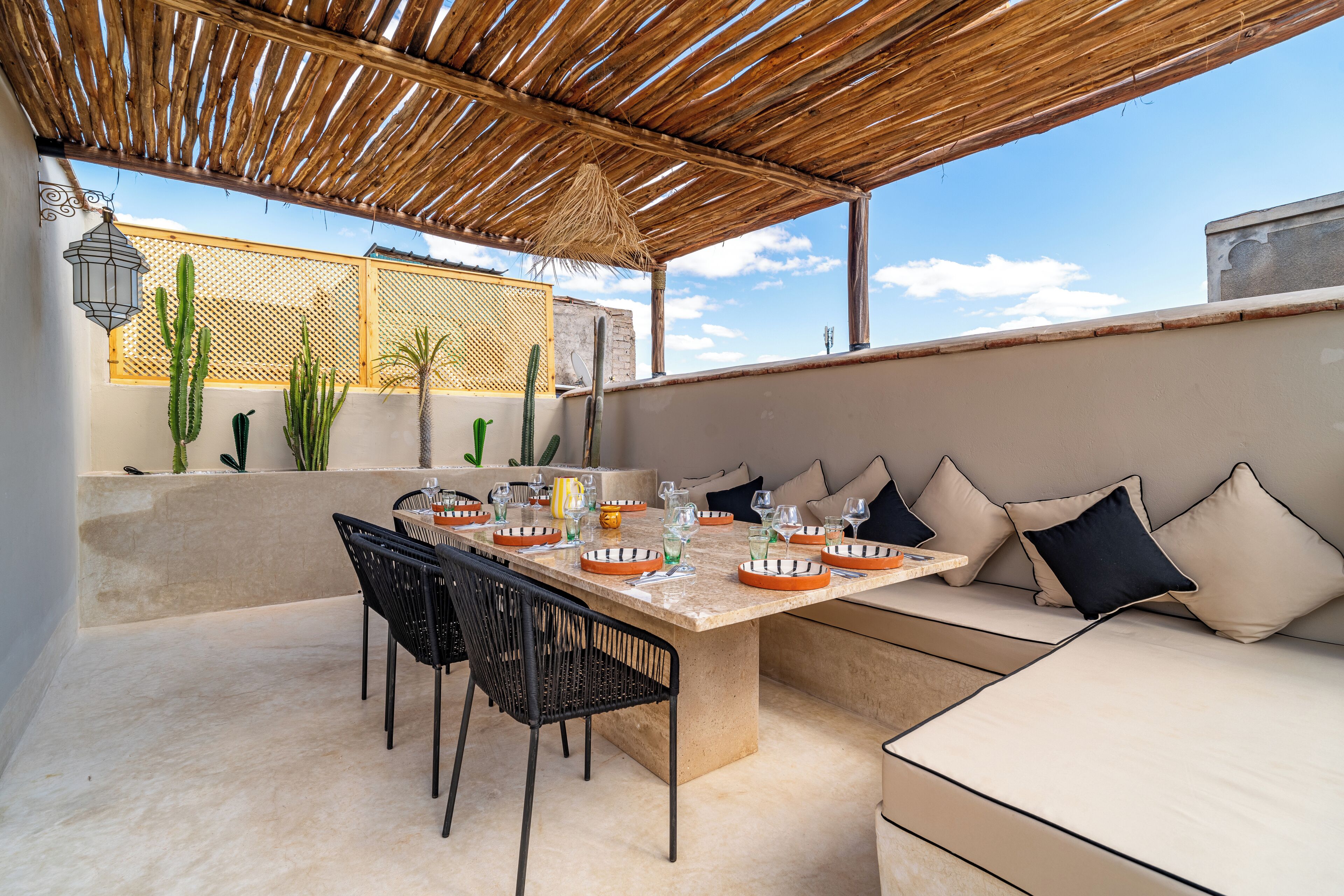 An outdoor dining area with a table, chairs, and a cushioned seating area under a thatched roof.