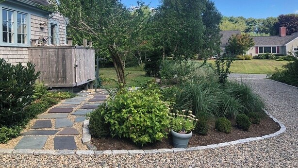 A house with a stone pathway leading to a wooden deck, surrounded by various shrubs and trees.