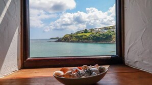 Ein Fenster mit Blick auf eine Küstenlandschaft, ein Holztisch und eine Schale Muscheln.