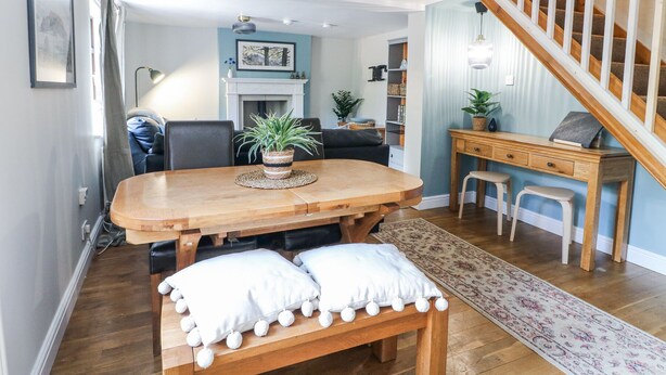 A dining area with a wooden table, two white cushioned benches, a woven placemat, and a potted plant. - Corner Cottage (Ashbourne)