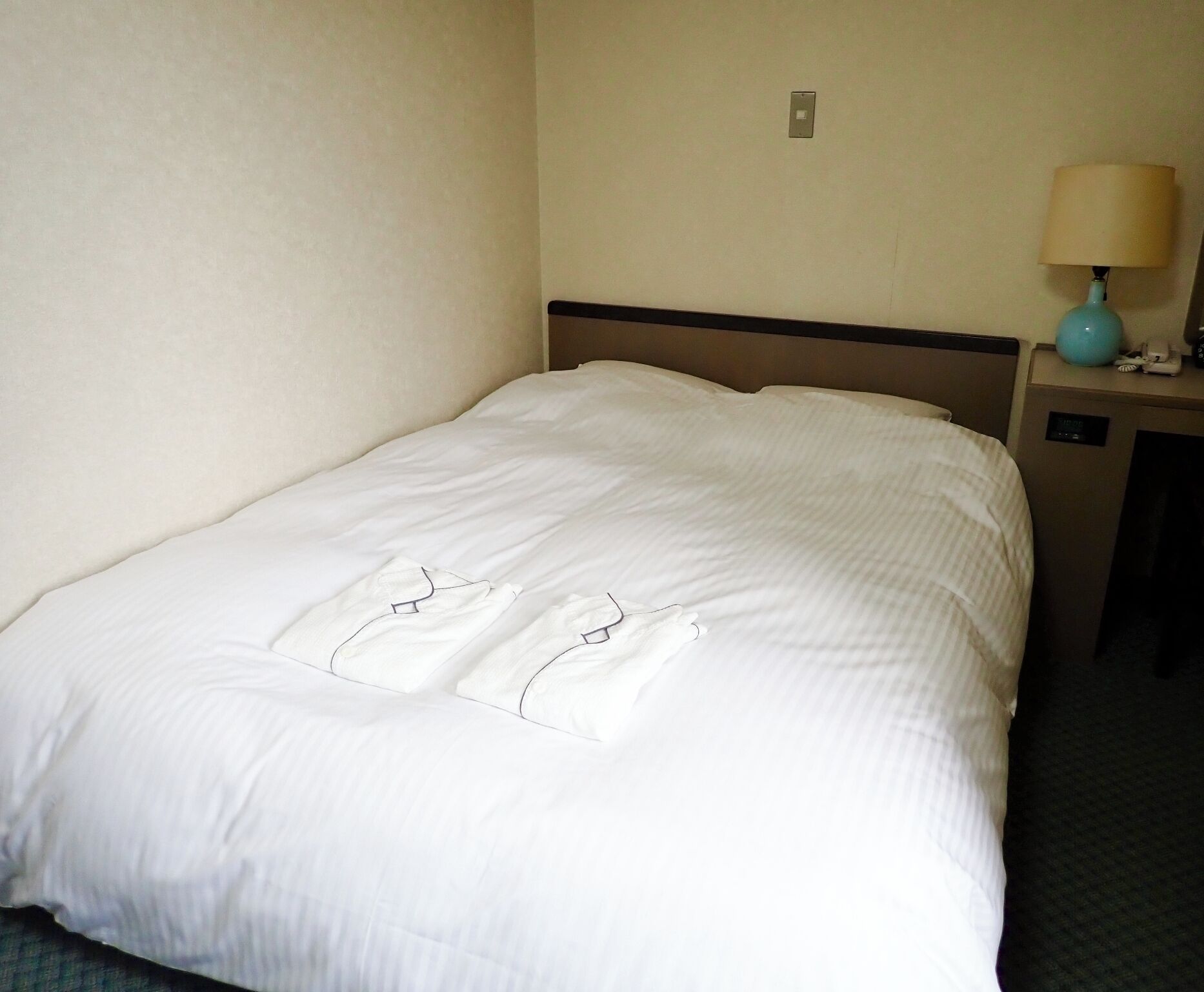 A neatly made bed with a white comforter and pillows, a bedside table with a lamp, and a wall socket.