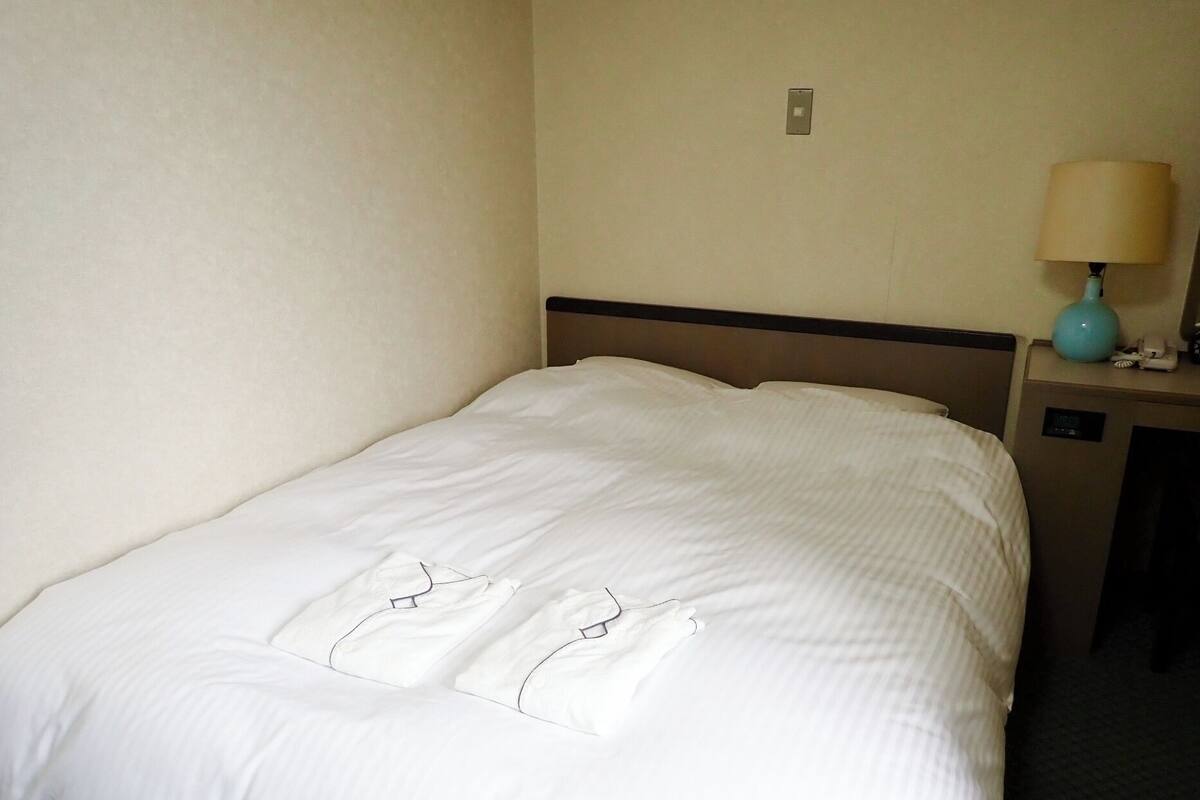 Una cama bien hecha con edredón blanco y almohadas, una mesita de noche con lámpara y un enchufe de pared.