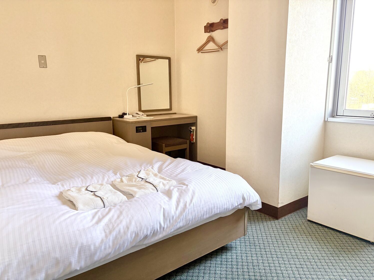 A hotel room with a bed, a bedside table with a lamp, a mirror, a small shelf, and a window.