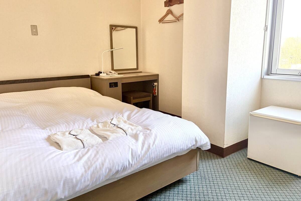 Una habitación de hotel con una cama, una mesita de noche con lámpara, un espejo, un estante pequeño y una ventana.