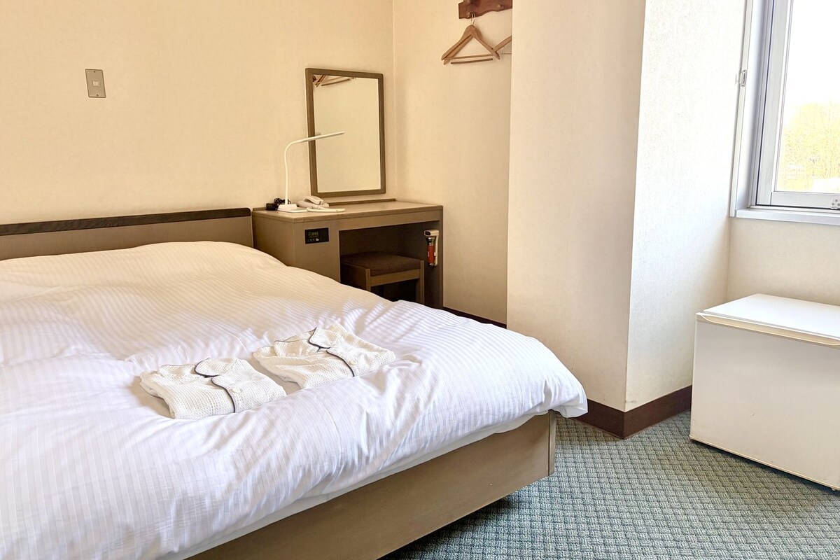 Una habitación de hotel con una cama, una mesita de noche con lámpara, un espejo, un estante pequeño y una ventana.