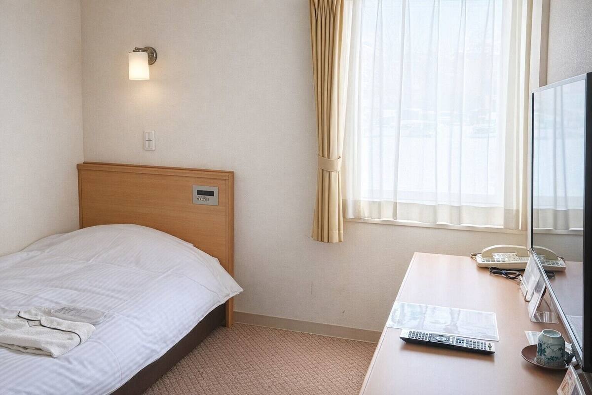 Habitación de hotel con cama, mesita de noche con teléfono y control remoto, ventana con cortinas e iluminación en la pared.