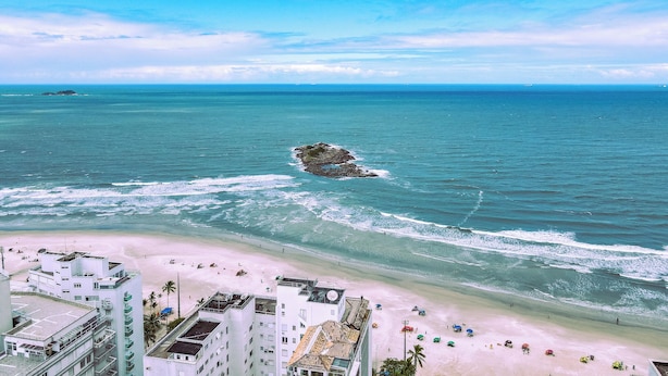 A coastal view with a sandy beach, ocean waves, and a rocky outcrop. - Beach Apartment - Air Conditioning and Barbecue (Guarujá)