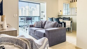 A modern living room with a grey sofa, a patterned throw blanket, and a dining area with a table and chairs. - Beach Apartment - Air Conditioning and Barbecue (Guarujá)