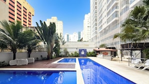 A swimming pool with two sections, surrounded by palm trees and a white building in the background. - Beach Apartment - Air Conditioning and Barbecue (Guarujá)