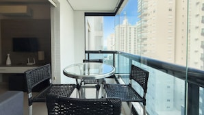 A balcony with a glass table and four wicker chairs, overlooking a cityscape with tall buildings. - Beach Apartment - Air Conditioning and Barbecue (Guarujá)