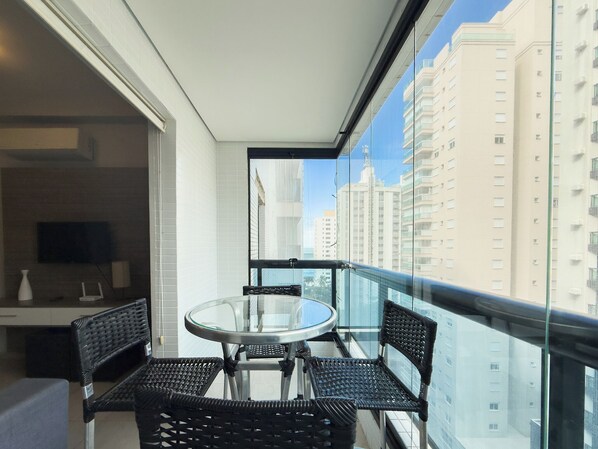 A balcony with a glass table and four wicker chairs, overlooking a cityscape with tall buildings.
