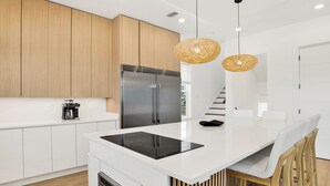 A modern kitchen with white cabinetry, a stainless steel refrigerator, a built-in oven, and a black induction cooktop. There are two wooden chairs with white cushions and a hanging woven light fixture. - Gulf Front Luxury | Private Heated, Cooled Pool | Elevator and EV Charging (Gulf Shores)