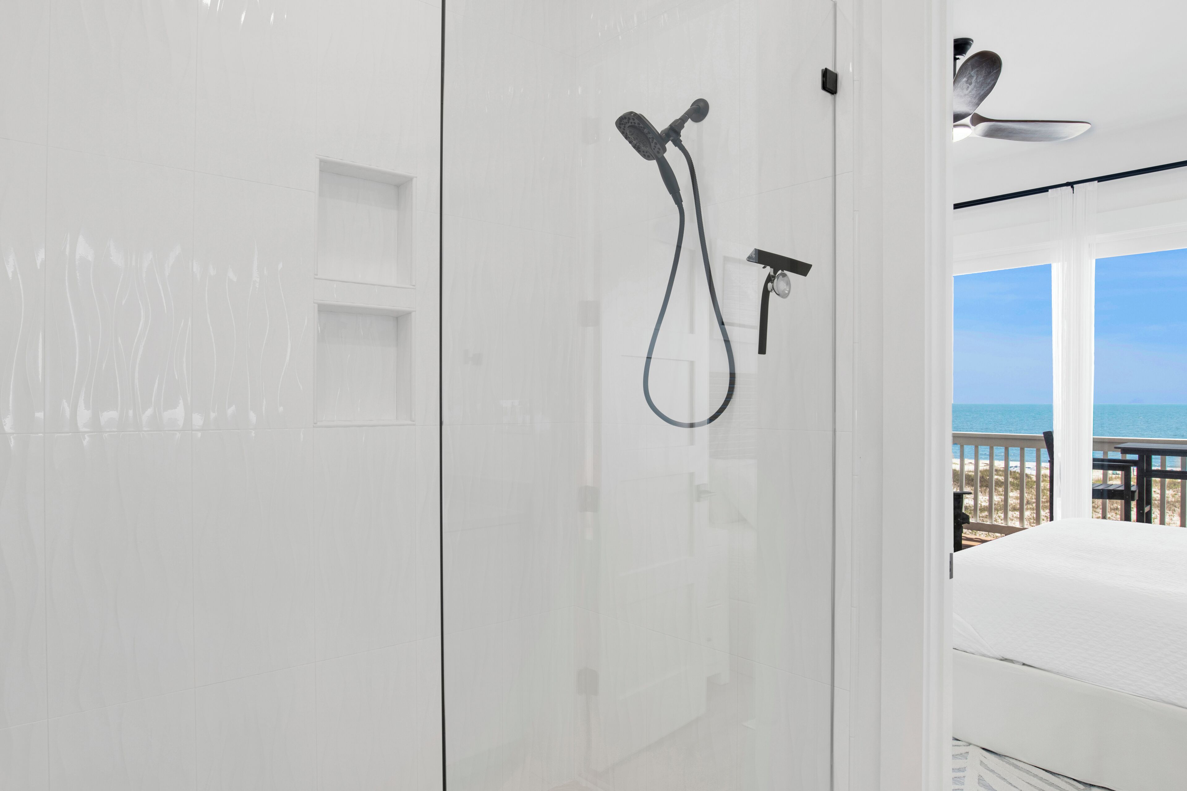 Une salle de bain moderne avec une douche à l’italienne en verre, un ventilateur de plafond et une vue sur la plage par une porte ouverte.