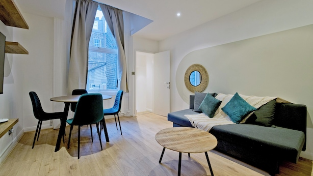A modern living room with a dining area, a round coffee table, a sofa with cushions, and a large window with curtains. - Imperial Queensborough Terrace (London)