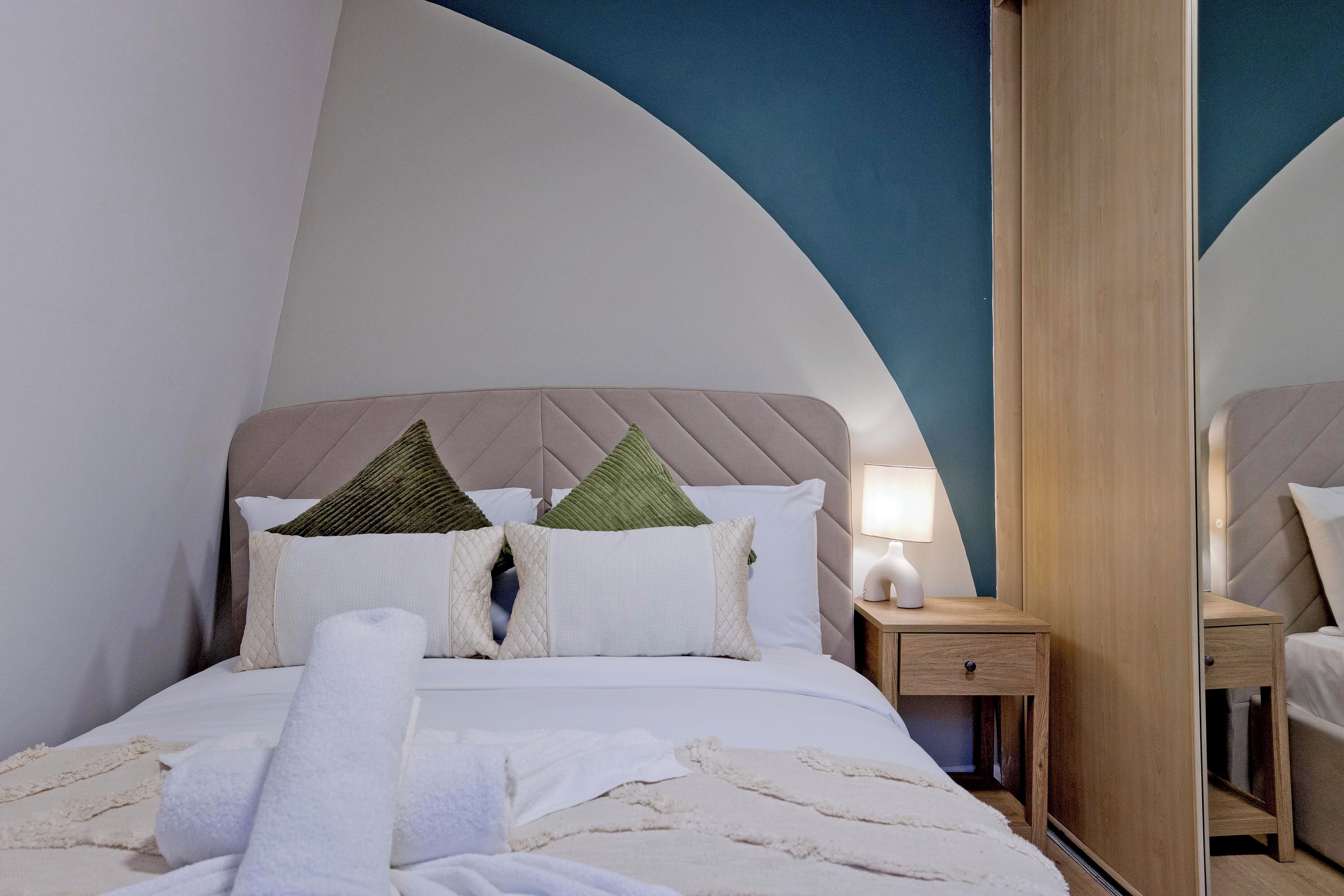 A modern bedroom with a curved ceiling, a bed with white linens and green pillows, a bedside table with a lamp, and a wooden wardrobe.