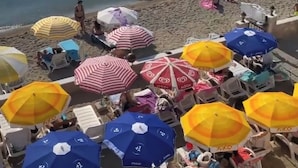 Ein Strand mit Sonnenschirmen und Liegestühlen, Booten im Wasser und badenden Personen.