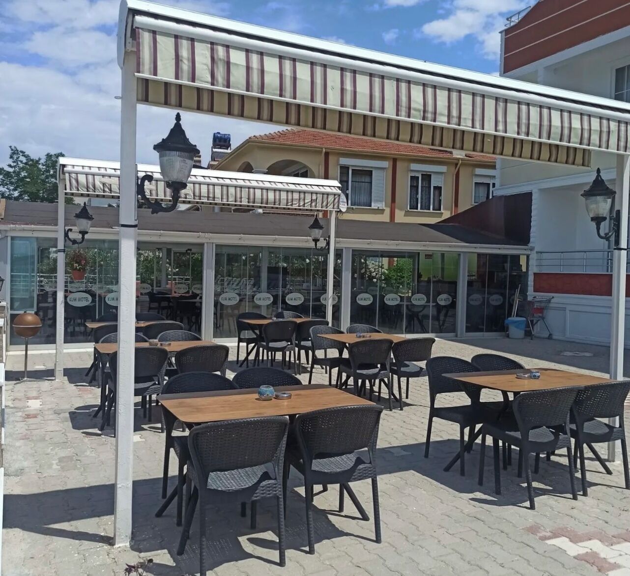 An outdoor cafe with tables and chairs, a covered seating area, and a building with a tiled roof in the background.