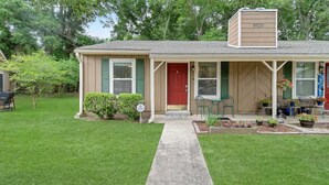 Une maison de plain-pied avec une porte rouge, un revêtement de couleur beige et un petit porche agrémenté de chaises vertes et de plantes en pot.