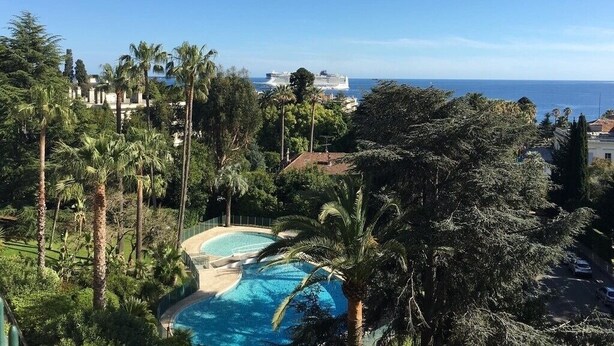 Une piscine entourée de palmiers, un ciel bleu azur et une vue sur l’océan, avec un navire de croisière qui se profile à l’horizon.