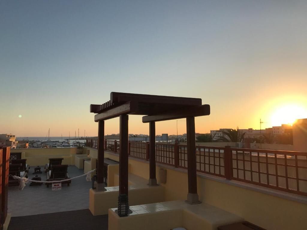 Una terraza en la azotea con una pérgola, zona de estar y vistas al atardecer sobre un puerto deportivo.
