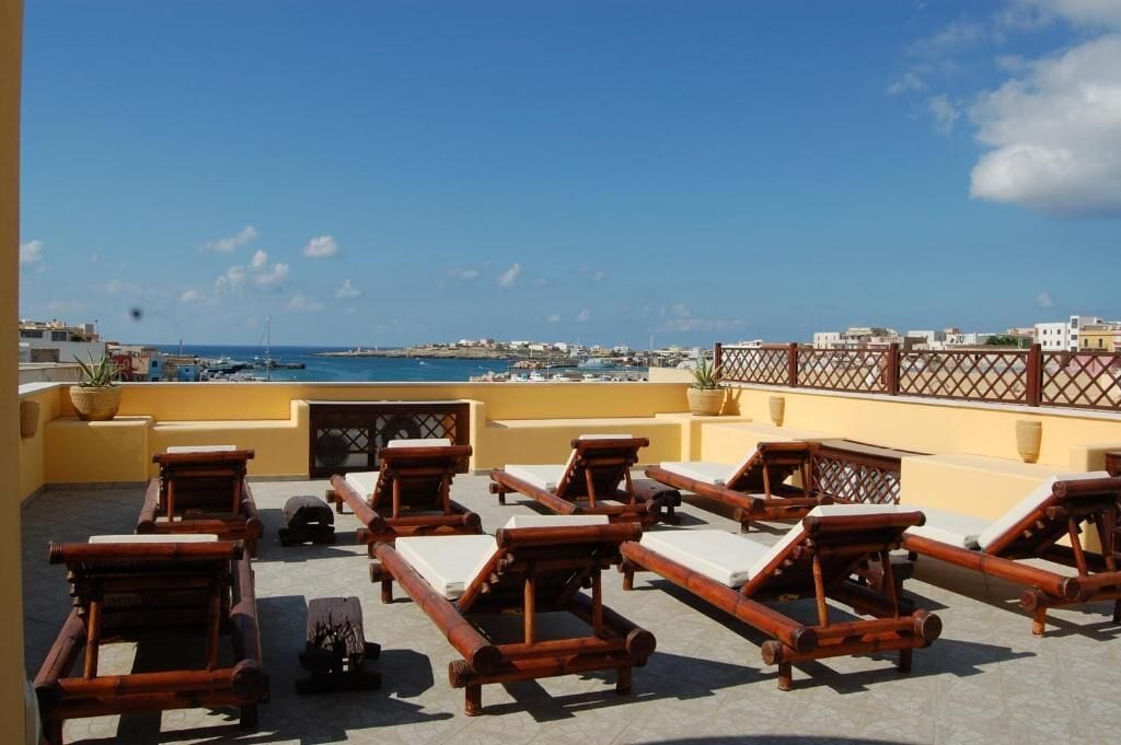 A rooftop terrace with wooden lounge chairs and a view of the sea and buildings.