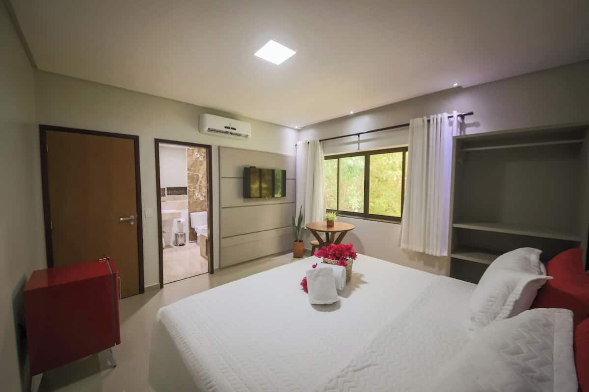 A hotel room with a bed, a red cabinet, a small table with flowers, a TV, and a window with curtains.