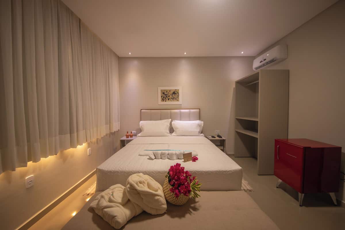 A hotel room with a neatly made bed, a bedside table with a lamp, a red cabinet, and a wall-mounted air conditioner.