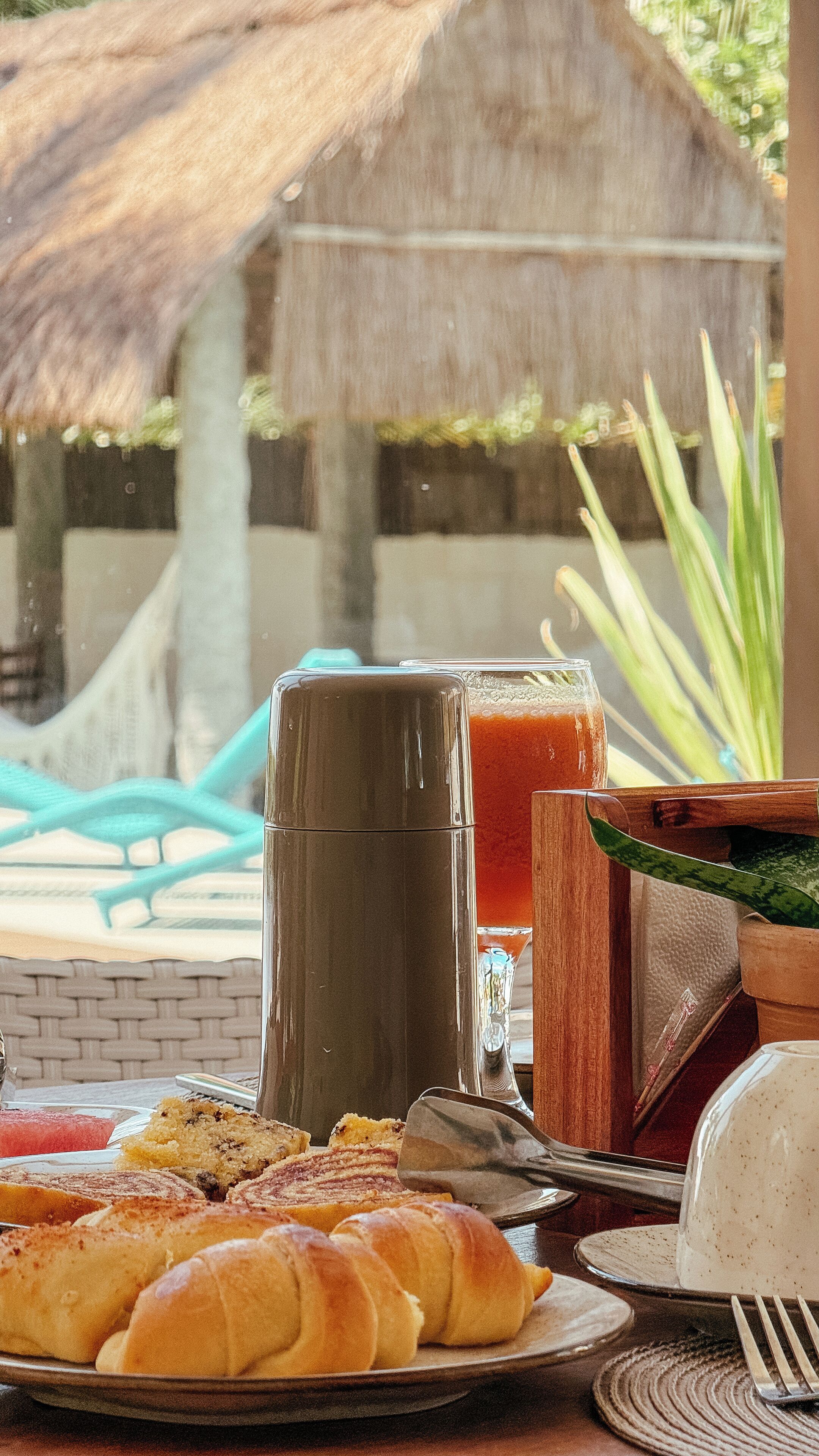 A breakfast table with croissants, a thermos, a glass of juice, and a wooden bowl.