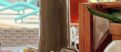 A breakfast table with croissants, a thermos, a glass of juice, and a wooden bowl.
