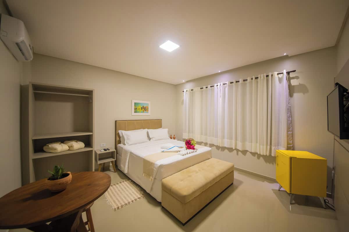 A hotel room with a bed, bedside tables, a small table with a plant, a bench, a yellow cabinet, and a window with curtains.