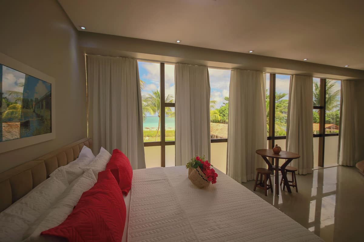 A hotel room with a large bed, a dining table with chairs, and a view of the beach.