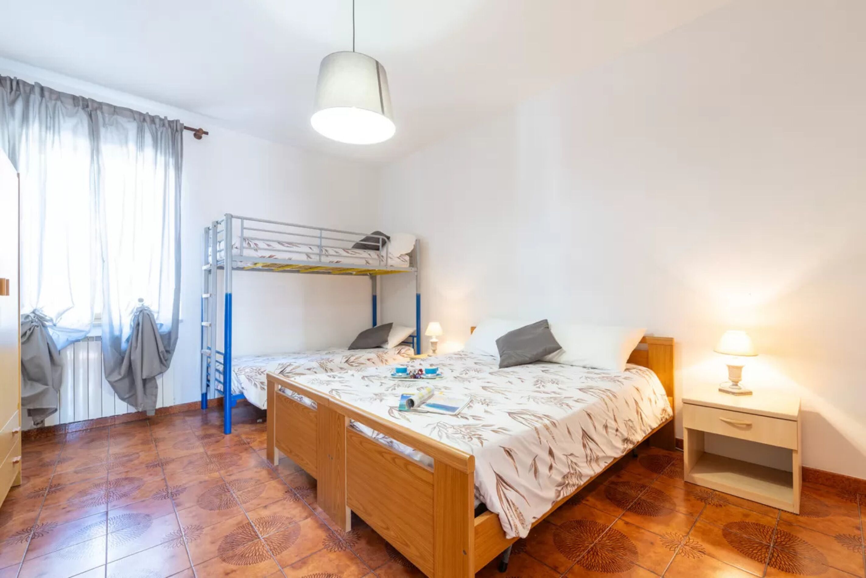 Ein Schlafzimmer mit Etagenbett, einem Einzelbett, einem Nachttisch, einer Lampe und einem Fenster mit Vorhängen.