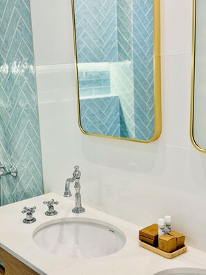 Une salle de bain avec un lavabo blanc, deux robinets et un miroir avec un cadre doré. On trouve des articles de toilette dans une boîte en bois posée sur le lavabo.
