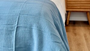 Ein ordentlich bezogenes Bett mit blauer Decke, einem Holzkopfstück und einem Nachttisch mit Lampe.