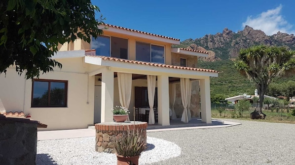 A modern two-story house with a patio area, a stone wall, and a view of mountains in the background. - CASA SULLA SPIAGGIA - ACCESSO DIRETTO ALLA SPIAGGIA DI SABBIA BIANCA (TERTENIA)