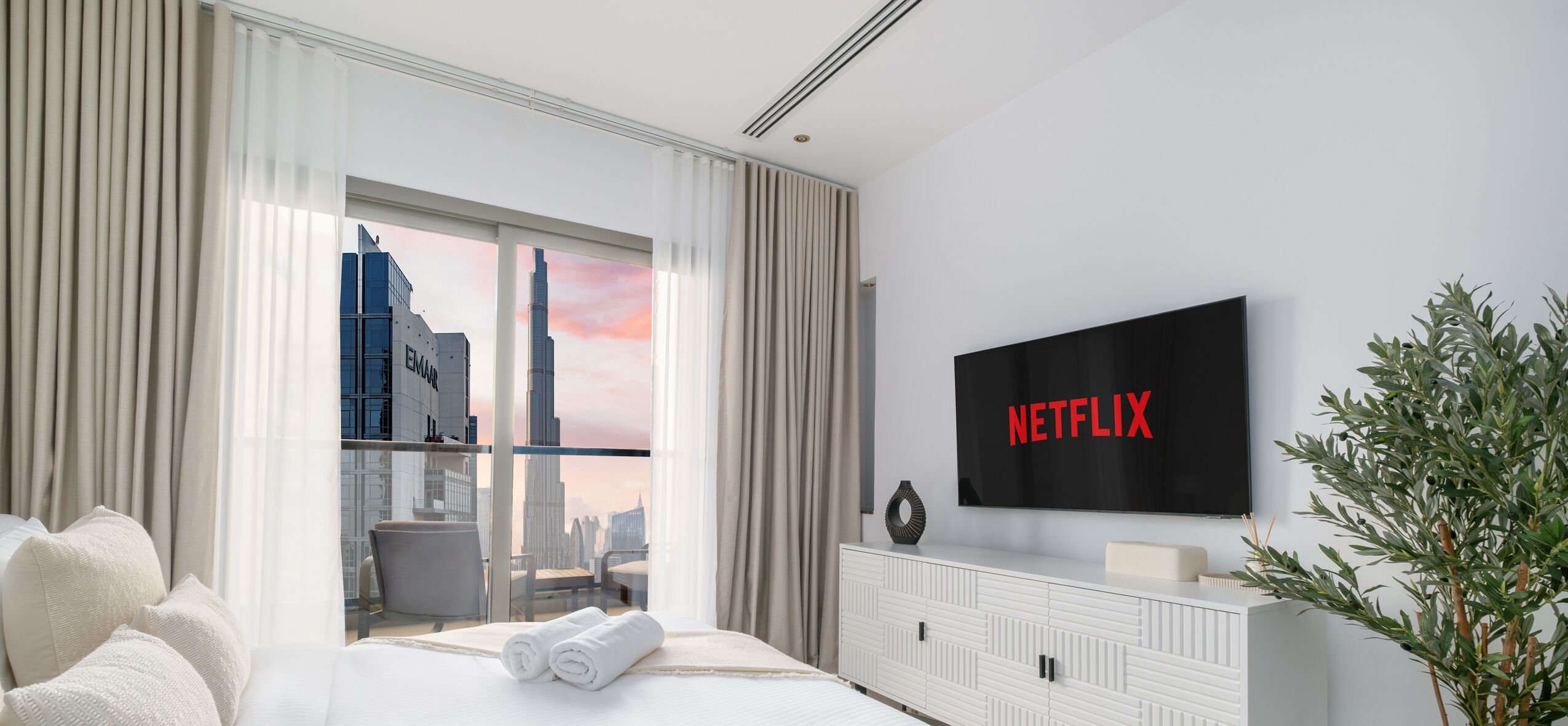 A modern hotel room with a large window offering a cityscape view, a flat-screen TV, and a neatly made bed.